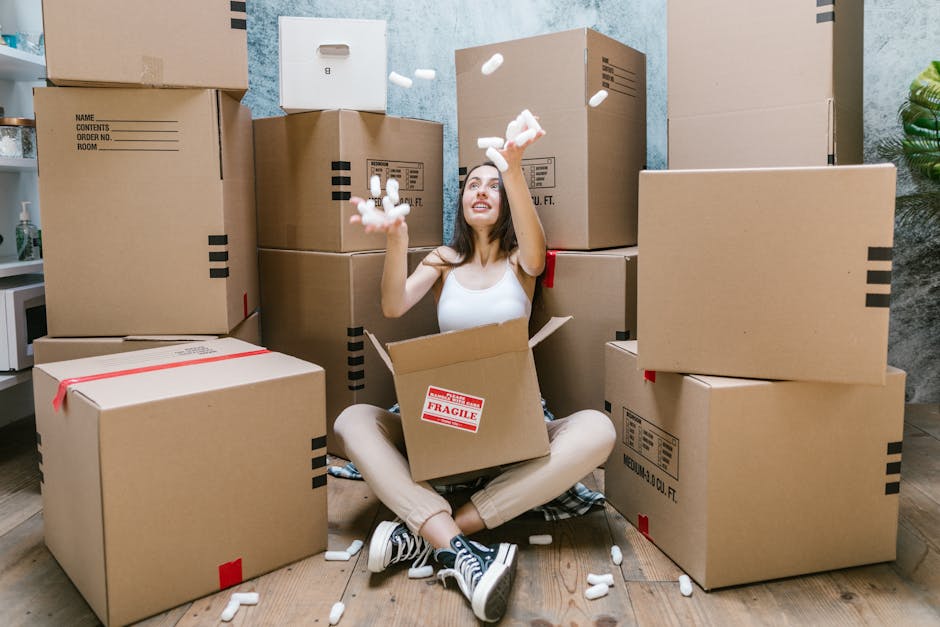A young woman smiling while holding an empty cardboard box labeled 'Fragile Glass' during a house removal in a room with a textured blue wall. She is standing among several packed cartons of various s