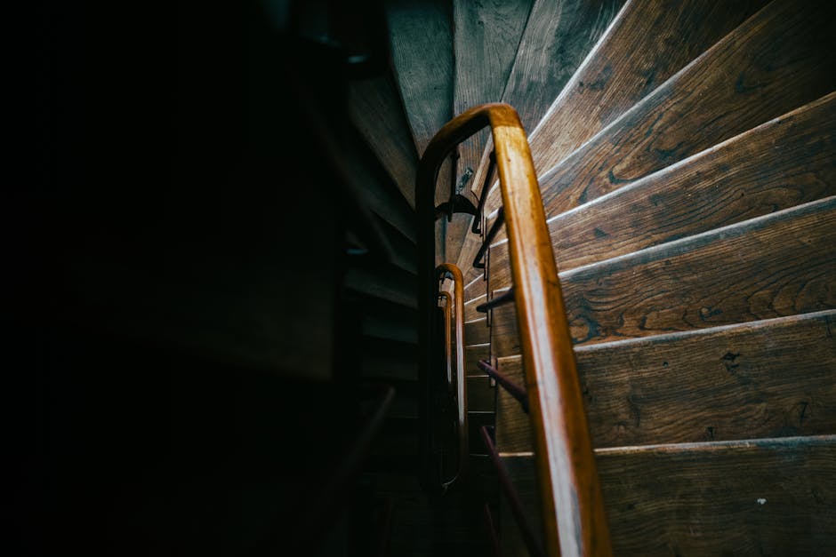 A narrow staircase inside a property with wooden steps and visible wear, leading upwards into a dark corridor. The staircase is framed by exposed wooden framing and located behind a doorway with a woo