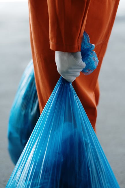Two workers dressed in orange high-visibility uniforms and white hats are engaged in a home relocation process during daytime. One worker is placing a wrapped and labeled cardboard box into the rear c